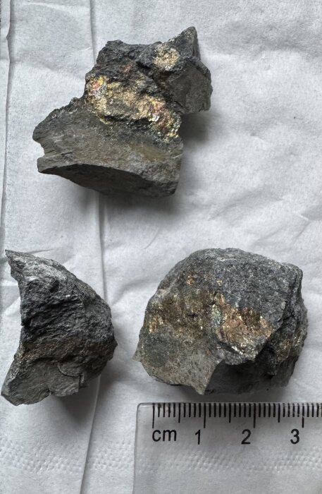 Tre stenprover med glittrande ytor, möjligen kvarts eller glimmerskiffer, ligger på papper med en linjal i bild för storleksreferens.