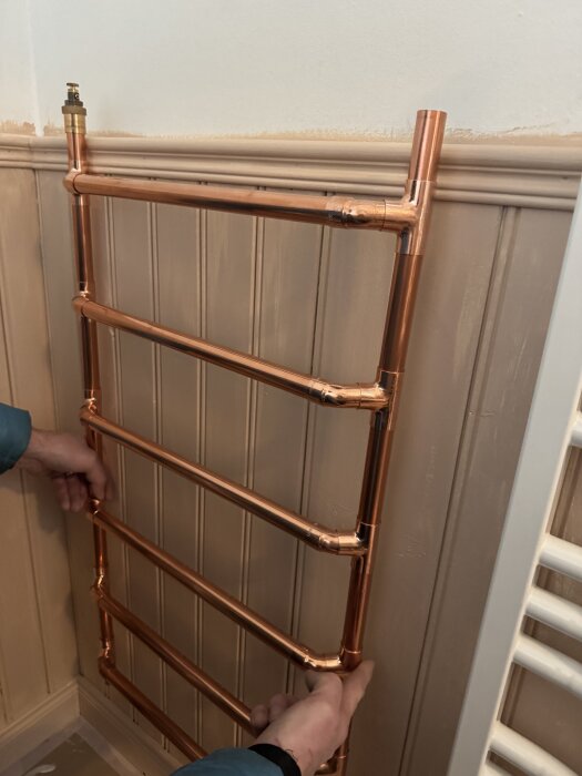 Handdukstork av kopparrör monteras mot träpanelvägg, händer justerar positionen.