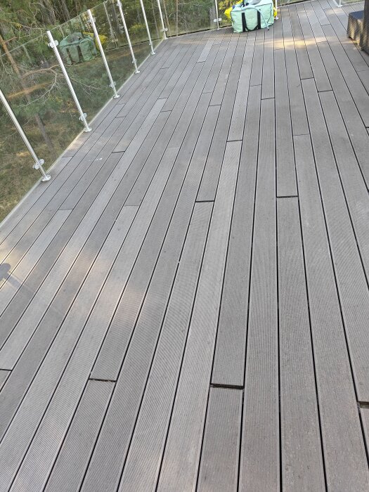 Komposittrall med springor på en altan med glasräcke. En stor gul förpackning syns i bakgrunden.