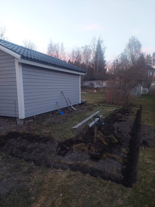 Grävd ut plats vid uthus för projekt med staket och kaninhus, rör och redskap syns. Skymning över tomt med träd och byggnader i bakgrunden.