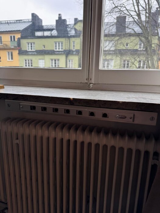 Fönster med karmventil ovanför ett element, med utsikt mot flerbostadshus och träd utanför. Ventilen är av en nyare modell.