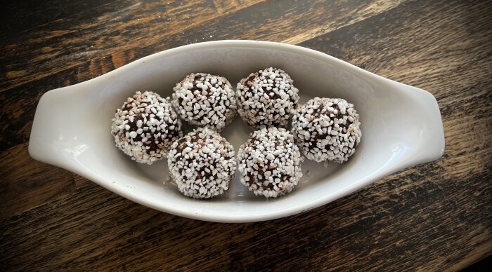 Chokladbollar täckta med pärlsocker i en vit oval skål på ett träbord.