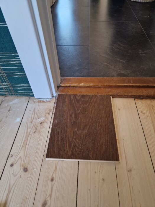 Parkettprov placerat mot tröskel och befintligt trägolv, dörrkarm och klinkergolv syns i bakgrunden.