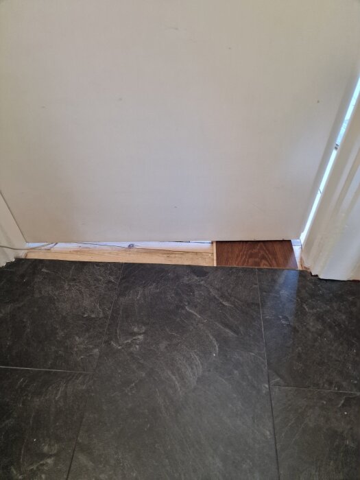 Dörröppning mellan två rum med olika golvbeläggning; mörk parkett synlig delvis under dörren, bredvid svart klinker i ett annat rum.