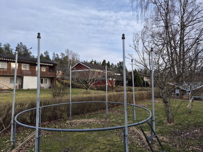 En cirkulär ram med åtta stolpar för ett skyddsnät på en gräsmatta, omgiven av träd och hus i bakgrunden.