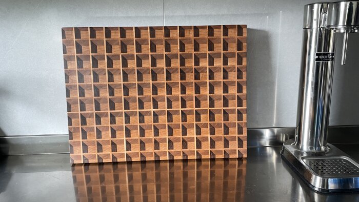 Skärbräda i trä med geometriskt mönster står lutad mot en metallvägg bredvid en vattenbehållare i rostfritt stål.