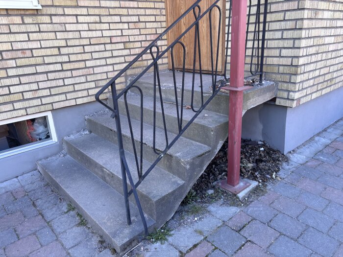 Trappsteg i betong med ett snett räcke bredvid röd byggstöd, bredvid ett tegelhus. En fönsterruta finns intill trappan.