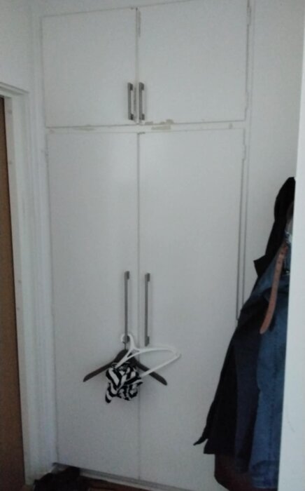 Vit garderob med dubbel dörr och metallhandtag, toppsektionen når inte taket. Klädhängare hänger på dörren.