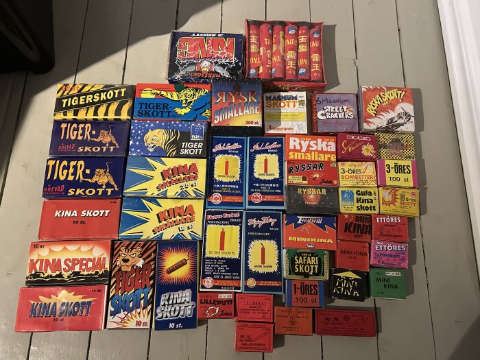 Gamla fyrverkeriförpackningar i olika färger och storlekar arrangerade på ett golv.
