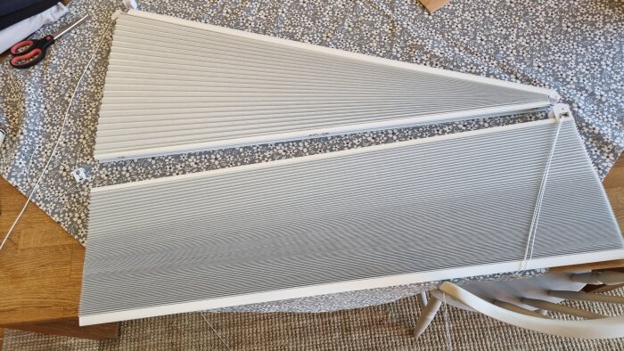 Gammal persienn med trasigt snöre liggande på ett mönstrat bord, med sax och snörstump bredvid.