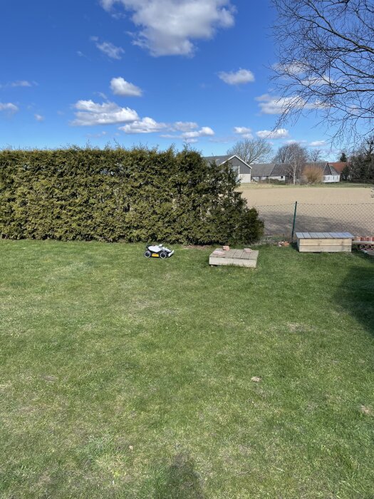 Gräsmatta med Luba Mini robotgräsklippare installerad, buskar i bakgrunden och klarblå himmel med moln.