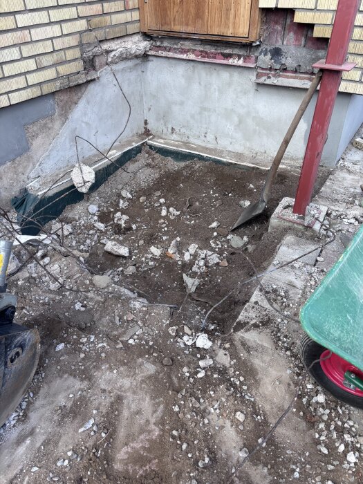 Grop med jord och stenar utanför en husvägg, del av ett bygge med spade och skottkärra synliga.
