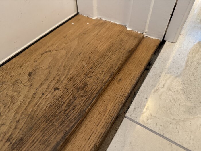 Tröskel i trä med slitage intill klinker, visar en glipa mellan golv och tröskel i renoverad entréhall.