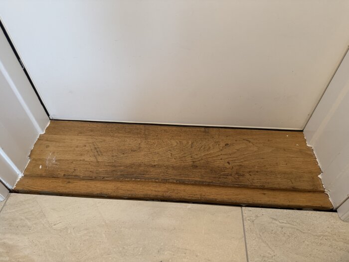 Tröskel i trä med glapp mellan golv och dörr. Omligger gränsar till ljust golv. Tröskeln är smutsig och ska slipas samt lackas.