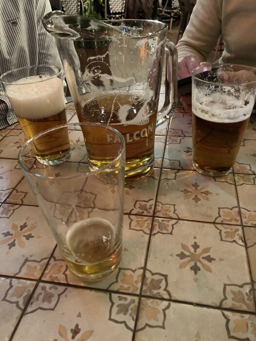 Fyra glas och en kanna öl på ett mönstrat bord, del av en kväll på lokal krog.