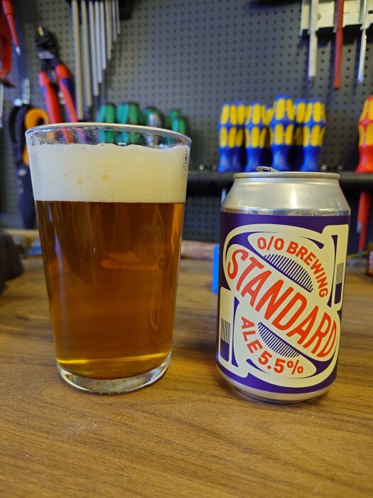 Ölglas med skummande ale och en burk märkt "O/O Brewing Standard Ale 5.5%" på ett bord i en verkstadsmiljö.