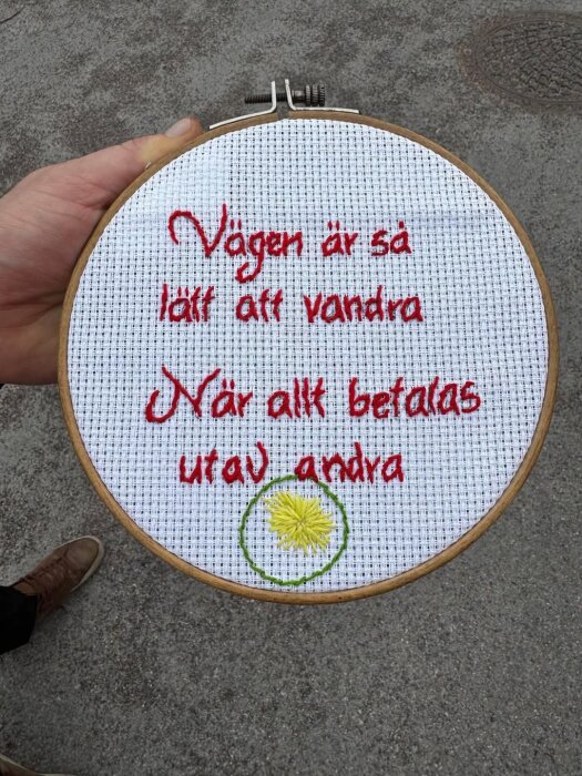 Broderad tavla med röd text: "Vägen är så lätt att vandra När allt betalas utav andra", samt en broderad gul blomma på vit bakgrund.