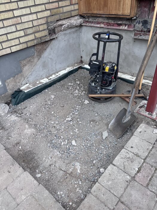 Byggarbetsplats vid en husfasad med jordyta och verktyg som spade och markvibrator på plats.