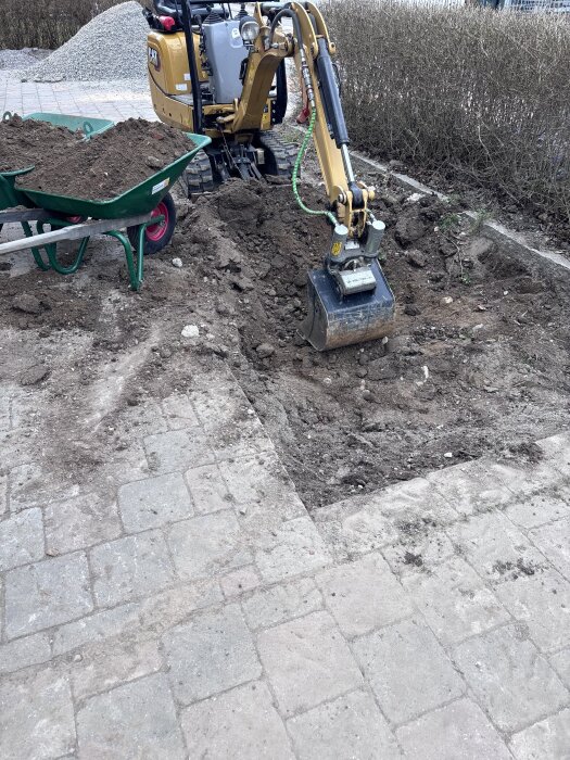 Minigrävare gräver i marken vid stenbelagd yta, med skottkärra fylld med jord bredvid.