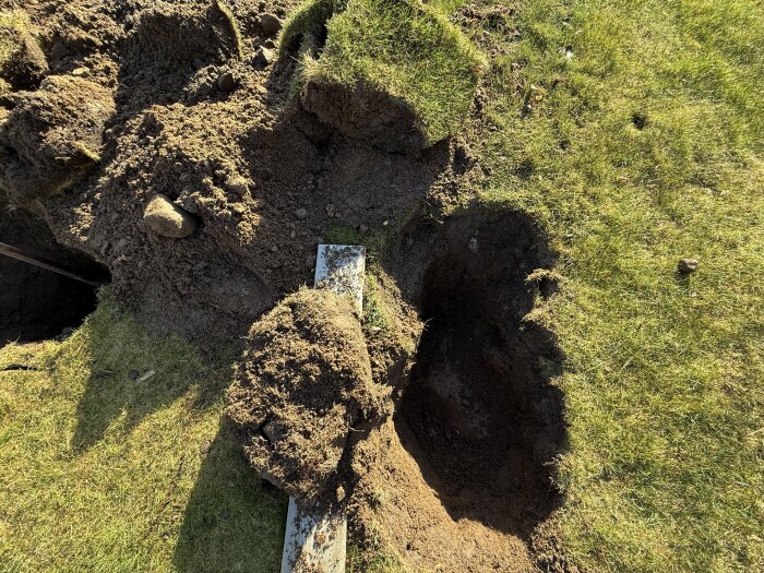 Utgrävt hål i gräsmatta med jordhögar runtom, del av förberedelse för bygge av altan runt pool.