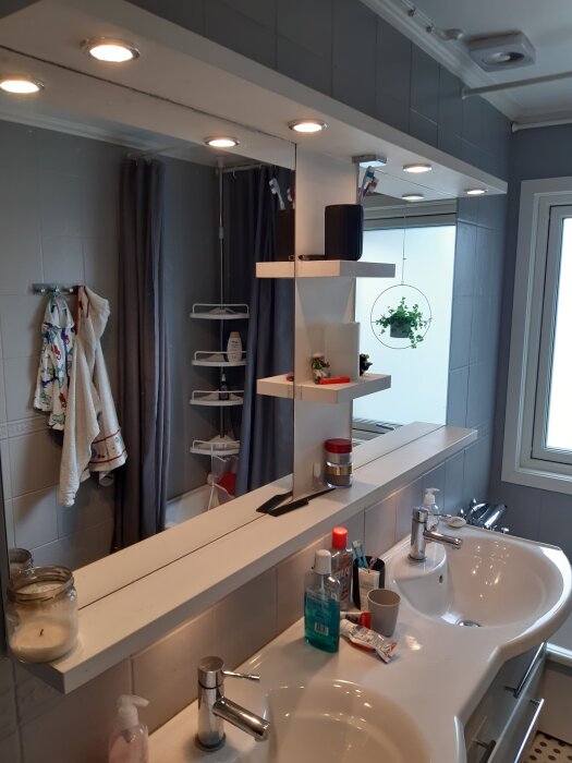 Badrum med dubbla handfat, spegel upplyst av spotlights och hylla ovan diskbänk. Tandborstar och hygienartiklar synliga.