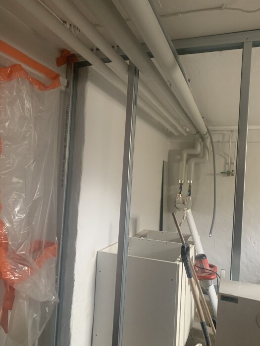 Rörledningar synliga i taket med stålreglar på väggen. Plast och byggutrustning synliga i rummet.
