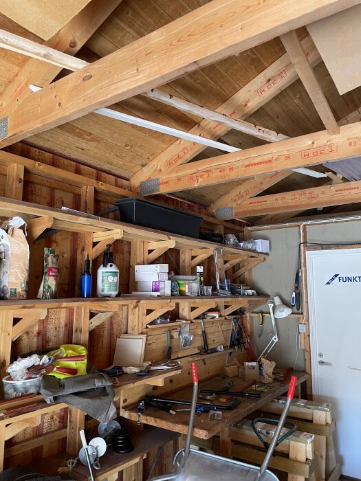 Kallgarage med öppna träväggar och tak, hyllor fyllda med olika verktyg och material för renovering och isolering.