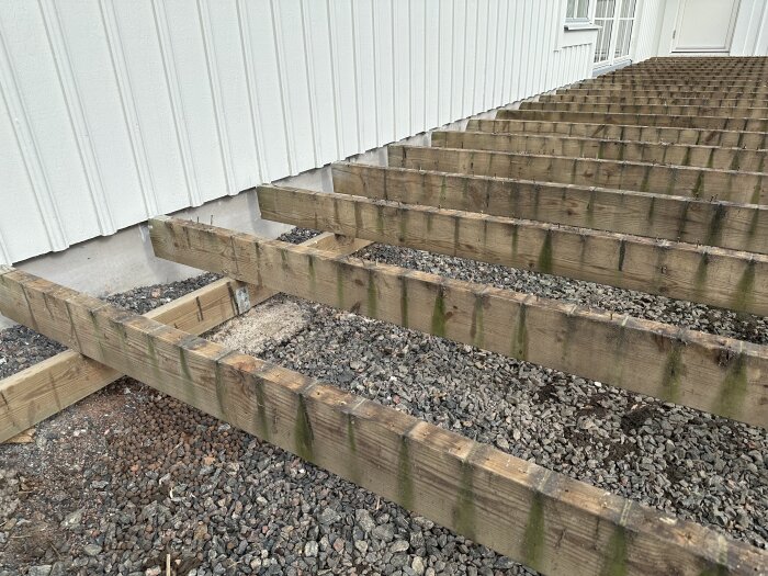 Rivna trallbrädor visar reglar och bärlinor på en altan byggd 1998, med grus under och närliggande husvägg med vit panel.