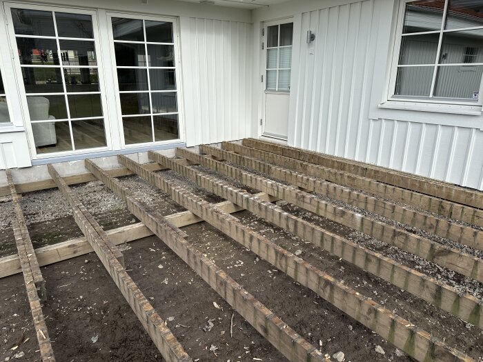 Altan med uppbruten trall visar synliga reglar och bärlinor mot en vit husvägg med stora fönster och en dörr. Grus och jord syns under.