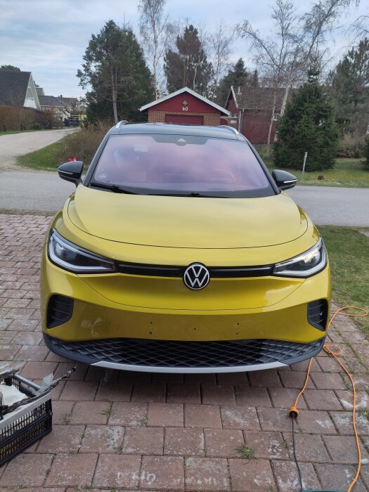 Gul Volkswagen utan registreringsskylt parkerad på uppfart, plastkorg och sladd synliga i förgrunden, tegeldäck och trähus i bakgrunden.