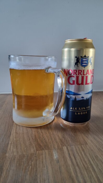 Ölmugg med gyllene dryck bredvid en burk Norrländska lageröl på ett träbord.
