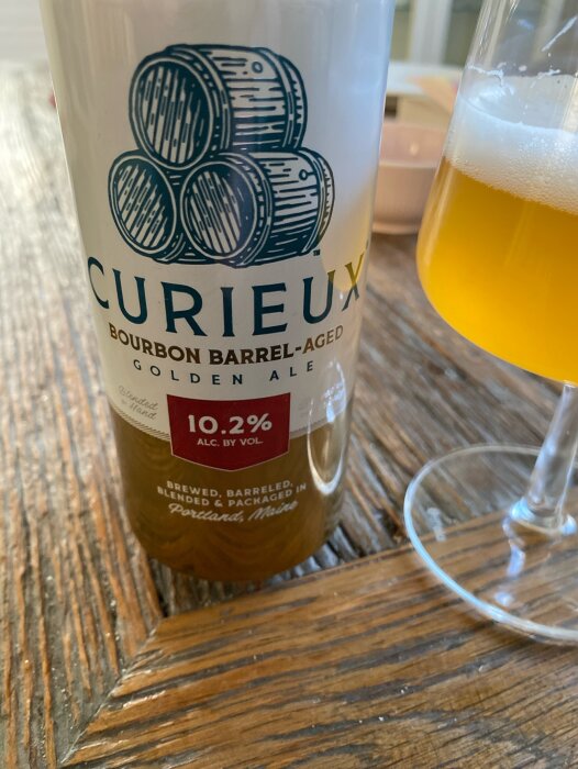 Flaska och glas med Curieux bourbon-fatlagrad golden ale från Allagash Brewing, 10.2% alkoholhalt, på träbord.