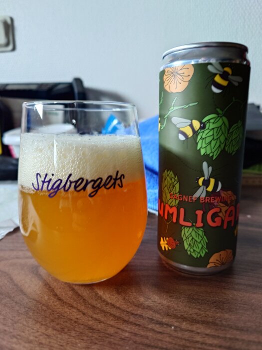 En ölburk märkt "Gagnef Brewing" med humle och bin, bredvid ett glas med ljus öl på ett bord.