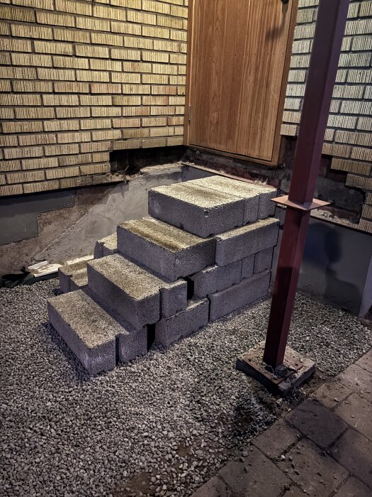 Nybyggd trappa av betongblock mot en tegelvägg, kringgärdad av makadam och en träport.