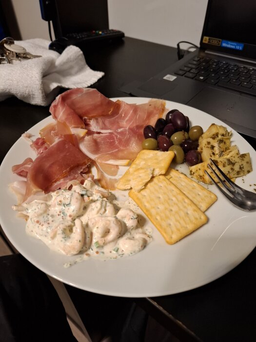 En tallrik med kallskuret, räkröra, kex och oliver på ett bord, bredvid en laptop och nycklar.