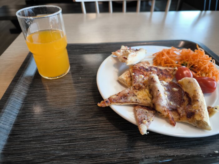Tallrik med pizzaslices, rivna morötter och tomater, bredvid ett glas apelsinläsk på ett bord.