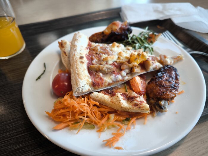 Tallrik med pizzaskivor, rivna morötter, tomat och kycklingvinge, serverad med dricka vid sidan.