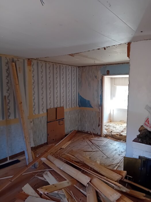 Rum under renovering med nedrivna träbitar på golvet, delvis rivet innertak och blottade väggar. Ljus kommer in genom en dörröppning.