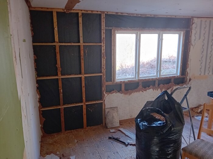 Renoveringsprojekt: Vägg med öppnad isolering, exponerade reglar, fönster, stege, stol och säck med byggmaterial i rum.
