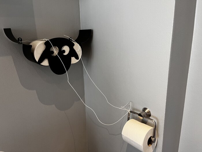 Toalettpappershållare i fårdesign ovanför en vanlig hållare med snöre för att påminna om byte av rulle när den är slut.