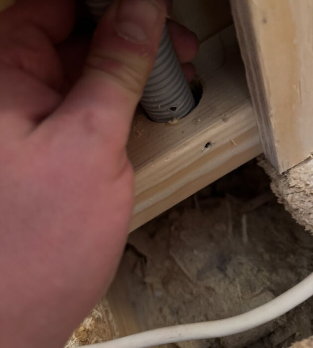 En hand håller ett skadat pex-rör i ett trångt utrymme, där det går genom ett hål i en träregel.