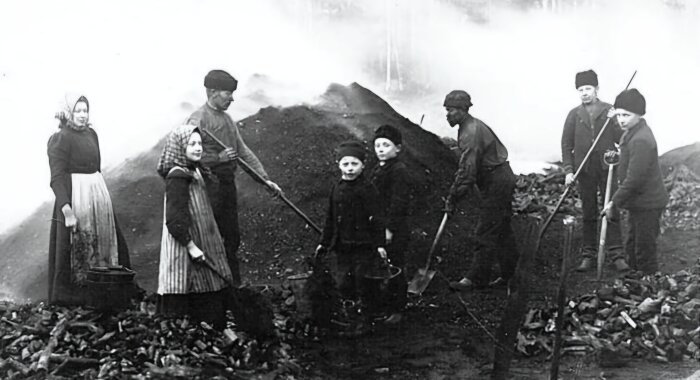 Personer arbetar vid en kolmila, iklädda traditionella kläder. Marken är täckt av träkol och rök syns i bakgrunden.