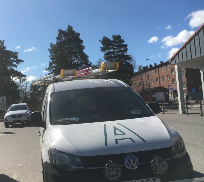 En vit skåpbil med stegar på taket står parkerad på en gata med träd och byggnader i bakgrunden.