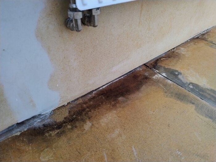Fuktskada på golv och vägg vid renovering, mörka fläckar längs väggens kant, nära rörkopplingar. Möjlig tidigare skada eller mögel antytt.