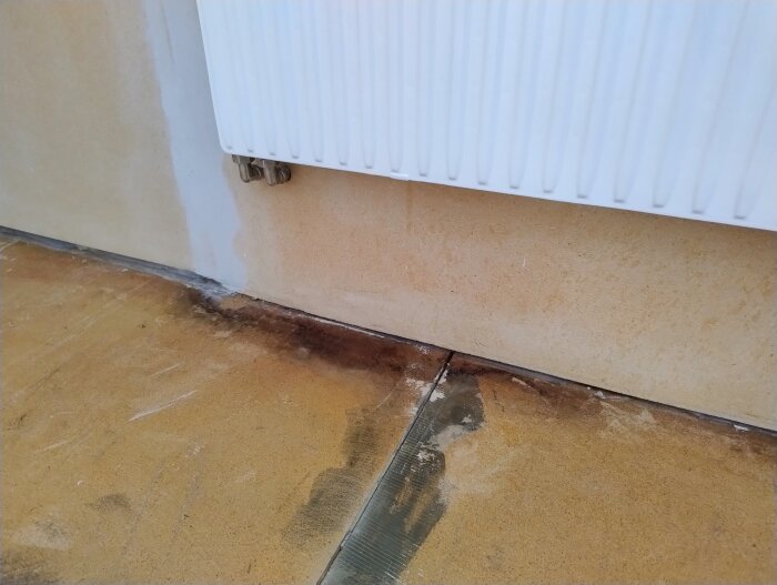 Fuktskadad undergolv vid radiator med mörka fläckar och sprickor, möjligen tidigare reparation, synlig vägg i sovrum under renovering.
