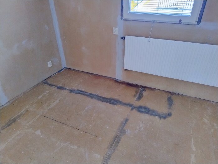 Fuktskada på sovrumsgolvet under plastmattan, med synliga mörka fläckar och sprickor vid golvbrädornas skarvar, bredvid en vägg med radiator.