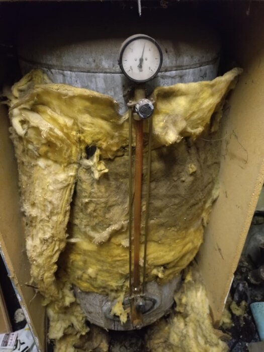 Hydrofor inpackad i gul isolering, med tryckmätare ovanpå och kopparrör anslutna. Tecken på åldrande och reparation behövs.