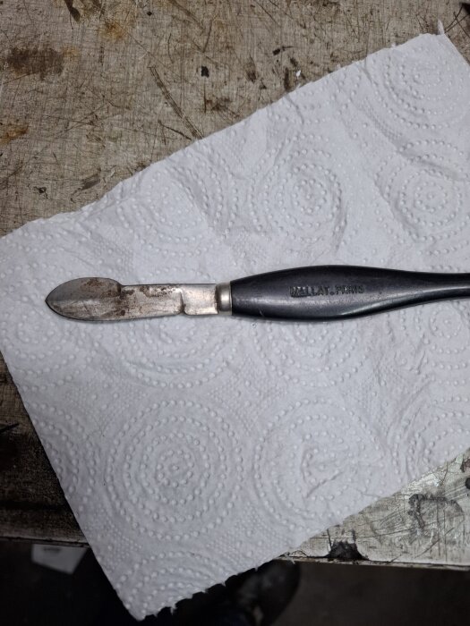 En okänd metallverktygsliknande föremål med en svart handtag, ligger på ett mönstrat papper på ett slitet bord.