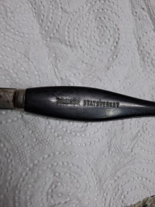 En metallisk föremål med texten "Tillhör Statsverket", liggandes på ett mönstrat papper.