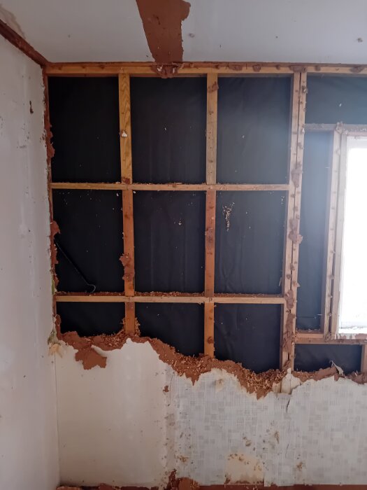 Inside view of a partially demolished wall showing wooden framing and black insulation. The structures appear unstable, revealing underlying construction materials.